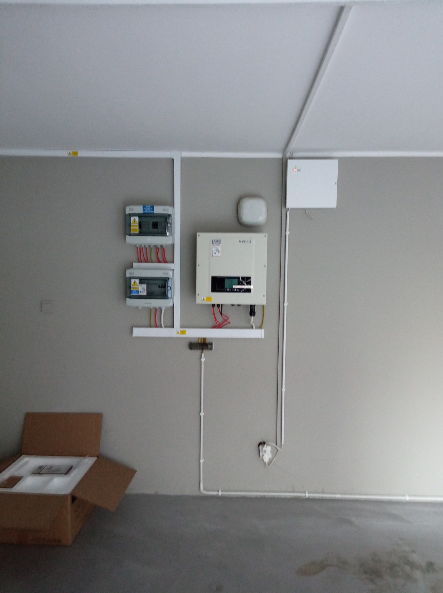 Panel fotowoltaiczny, rozdzielnice i okablowanie zamontowane na ścianie w garażu. Częściowo rozpakowane elementy instalacji leżą w kartonie na podłodze.