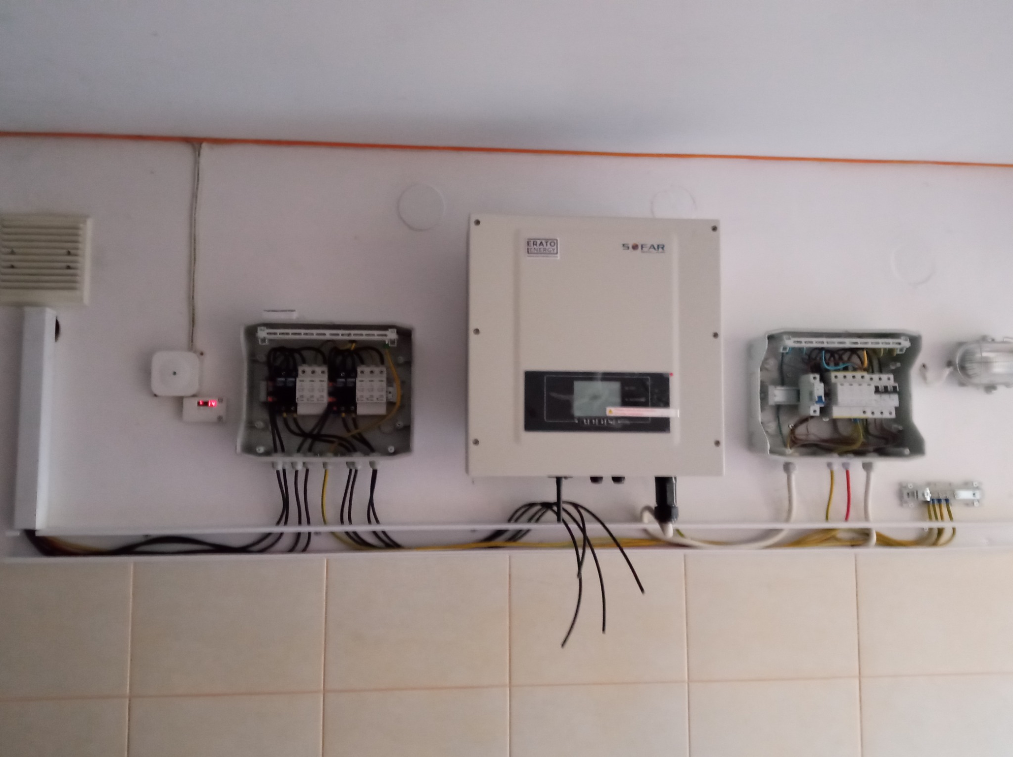 Rozdzielnice elektryczne i falownik SOFAR zamontowane na ścianie z kafelkami, widoczne przewody i elementy instalacji. Białe ściany i pomarańczowa rurka na suficie.