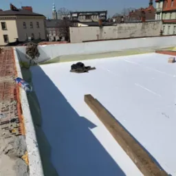 Białe płyty izolacyjne na dachu budynku w trakcie prac. Widoczne narzędzia i materiały budowlane. W tle zabudowa miejska.