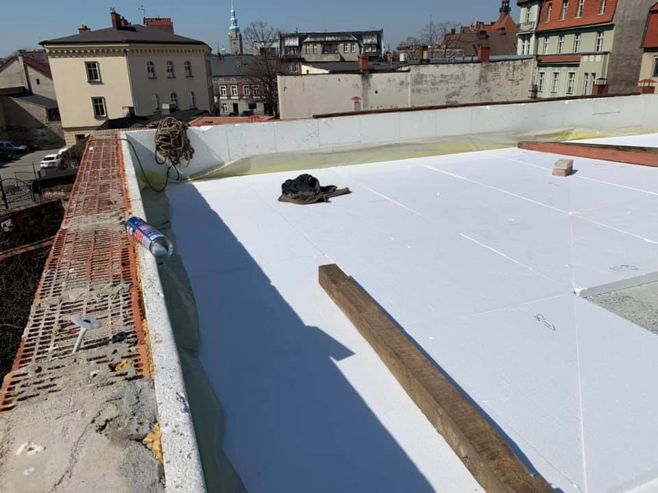 Białe płyty izolacyjne na dachu budynku w trakcie prac. Widoczne narzędzia i materiały budowlane. W tle zabudowa miejska.