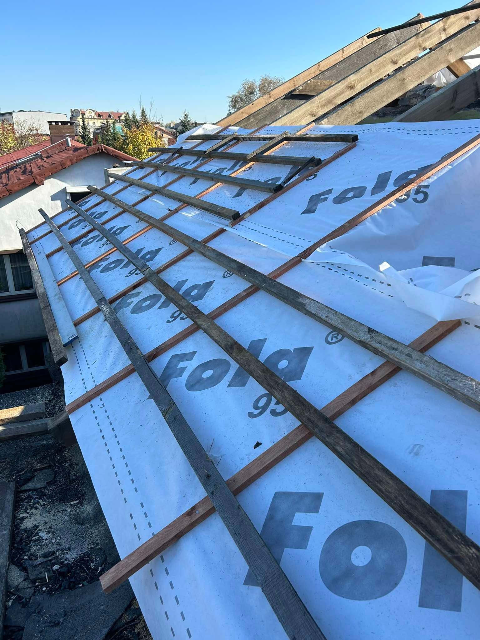 Dach w trakcie krycia: biała membrana dachowa z nadrukiem, drewniane łaty, widoczna konstrukcja więźby dachowej, fragment dachu sąsiedniego budynku i błękitne niebo.