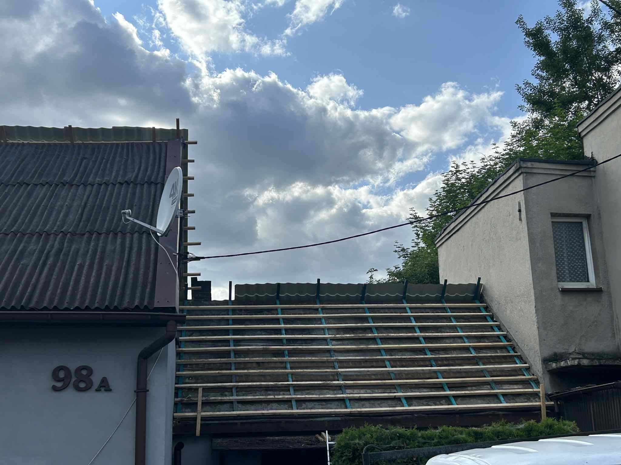 Remont dachu domu z numerem 98A, widoczne drewniane łaty i stara dachówka. W tle błękitne niebo z chmurami.