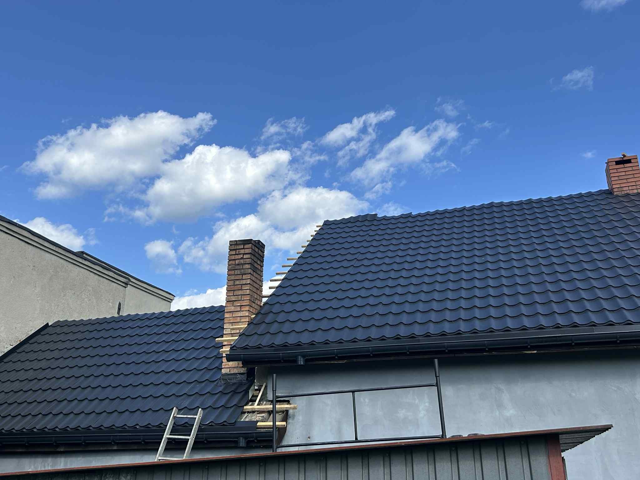 Czarny dach z blachodachówki, widoczny komin z cegły, drabina oparta o ścianę. Błękitne niebo z chmurami w tle. Widok z dołu na fragment dachu.
