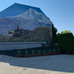 LEONID PODOLYAK MOREMURALS - Duży mural na ścianie budynku przedstawiający górski krajobraz z zamkiem na tle błękitnego nieba, widok z poziomu brukowanego podjazdu z rabatami kwiatowymi.
