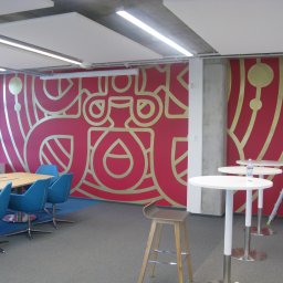 LEONID PODOLYAK MOREMURALS - Ściana w biurze z czerwonym tłem i złotym, abstrakcyjnym wzorem geometrycznym, w tle biurko z niebieskimi krzesłami, wysokie białe stoliki i drabina.