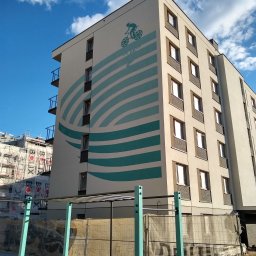 LEONID PODOLYAK MOREMURALS - Wielopiętrowy budynek z nowoczesną elewacją, ozdobiony artystycznym malunkiem przedstawiającym rowerzystę i abstrakcyjne turkusowe pasy, widoczny z poziomu gruntu, z elementami placu budowy w tle.