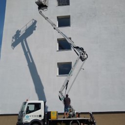 LEONID PODOLYAK MOREMURALS - Pracownik w podnośniku koszowym na tle białej elewacji budynku z napisem, cień podnośnika na ścianie, na dole samochód firmy Jartech i rabata z różami.