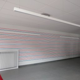 LEONID PODOLYAK MOREMURALS - Wnętrze biura z pomalowaną ścianą w poziome, naprzemienne pasy koloru czerwonego i niebieskiego. Widoczny biały grzejnik, sufit kasetonowy z lampą liniową i kratką wentylacyjną oraz drabina oparta...