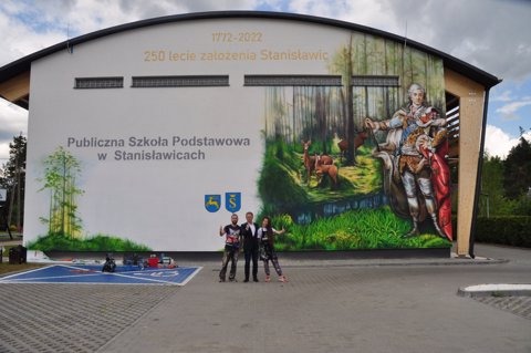 Duży mural na ścianie szkoły podstawowej w Stanisławicach przedstawiający postać historyczną w otoczeniu lasu i zwierząt, z napisem upamiętniającym 250-lecie założenia, oraz grupą trzech osób...