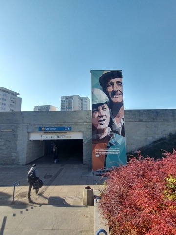 Billboard z artystycznym portretem dwóch mężczyzn przy wejściu do stacji metra Ursynów w Warszawie, z przechodzącą osobą w tle.