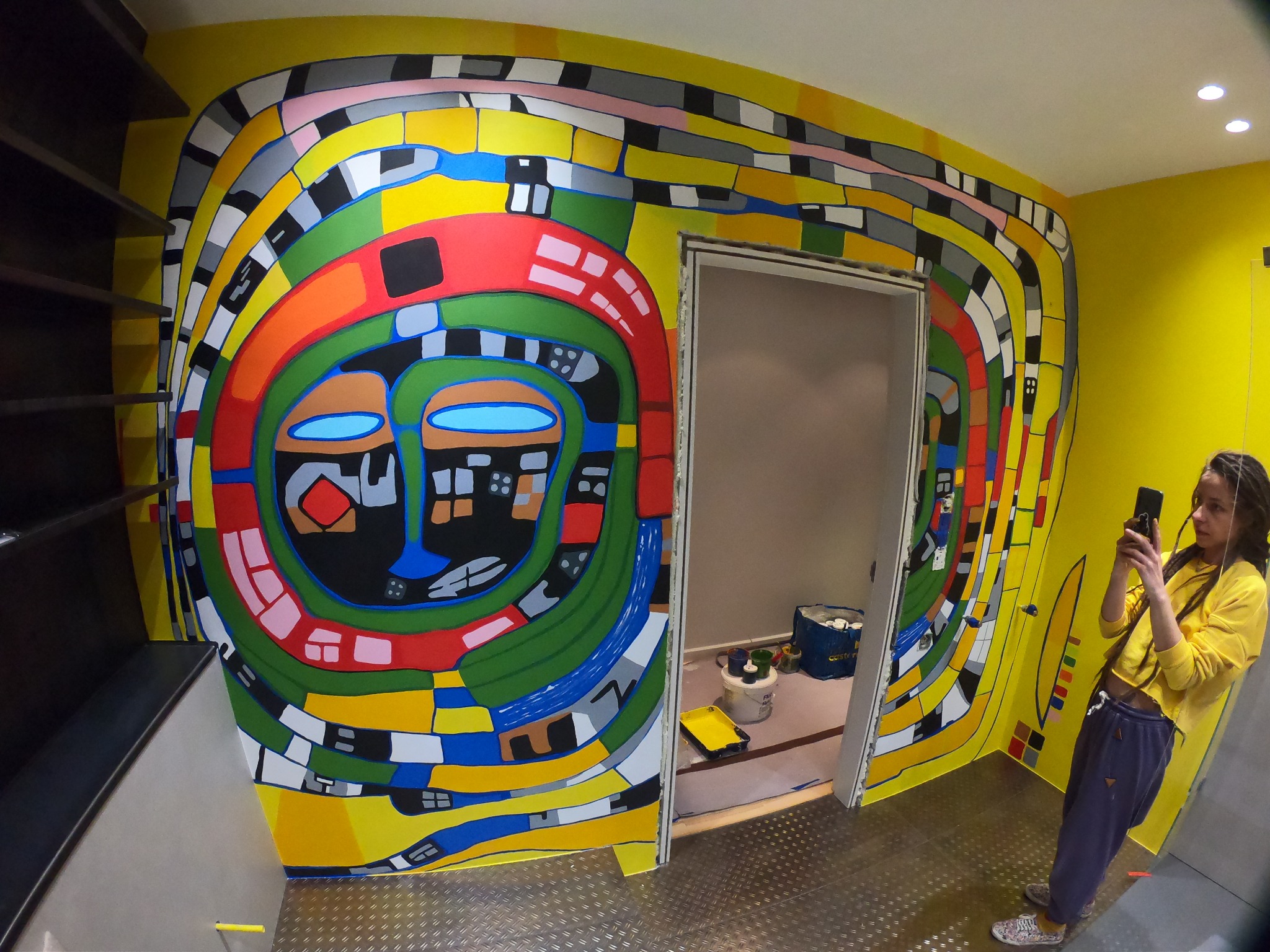 Kobieta fotografuje telefonem świeżo pomalowaną ścianę z abstrakcyjnym, kolorowym wzorem przypominającym twarz, widoczne pędzle i farby w otwartych pojemnikach w sąsiednim pomieszczeniu.