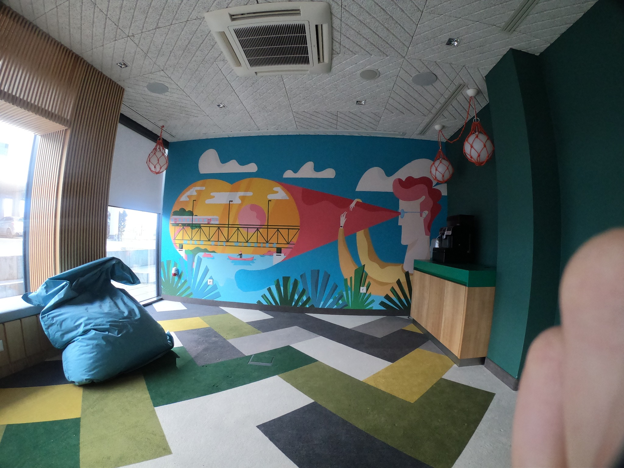 Kolorowe, artystyczne malowidło ścienne w biurze w Warszawie, przedstawiające abstrakcyjny krajobraz miejski z elementami natury, oświetlone naturalnym światłem wpadającym przez okno. W tle...