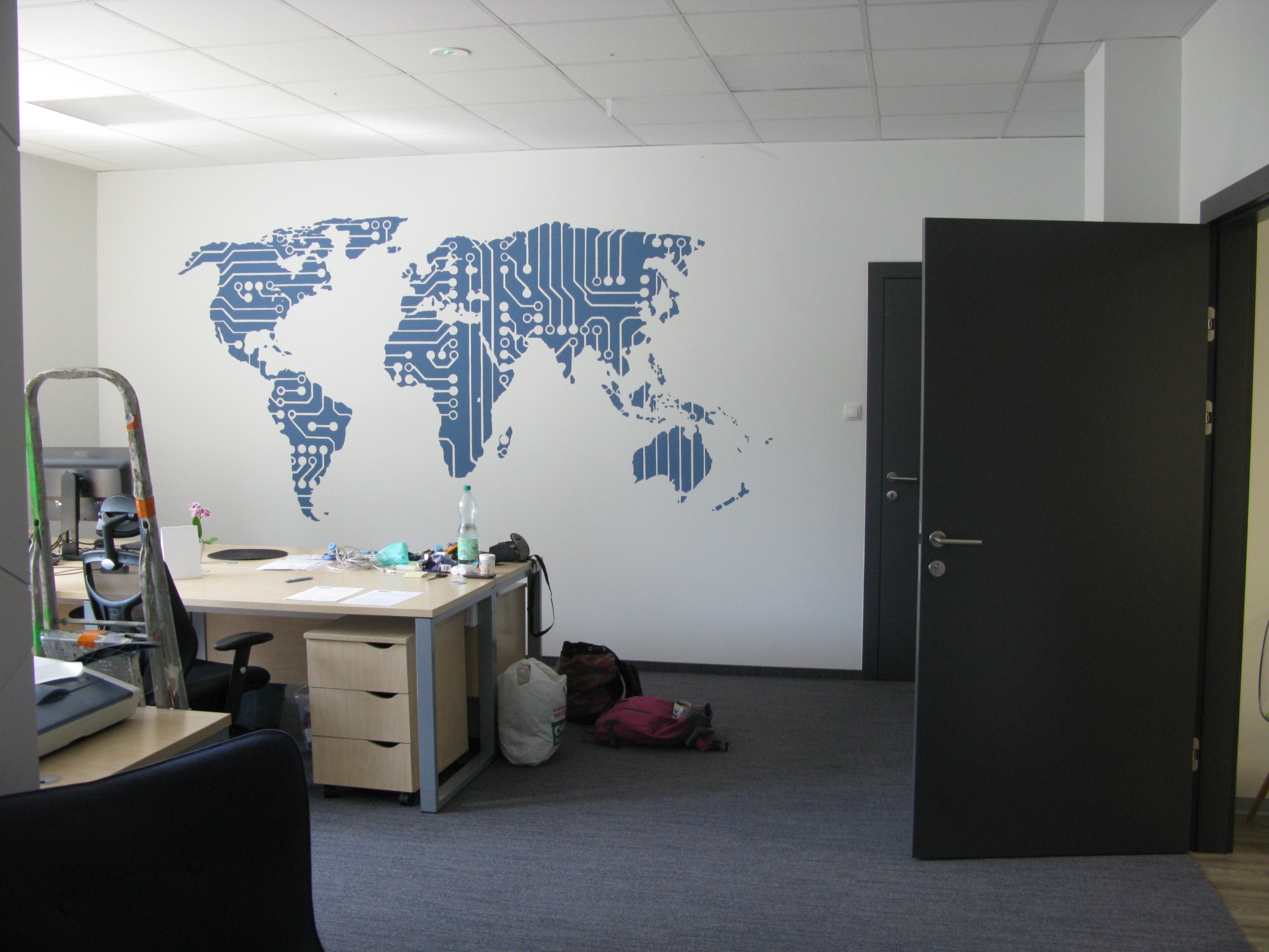 Biuro z biurkiem, krzesłem, torbami na podłodze i dekoracyjną mapą świata w formie układu scalonego na ścianie.