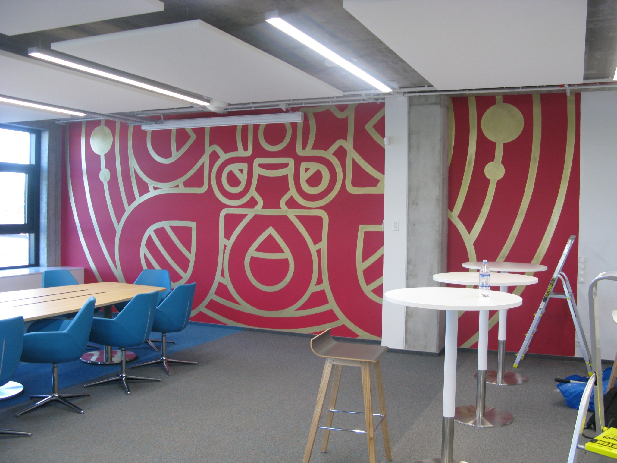 Ściana w biurze z czerwonym tłem i złotym, abstrakcyjnym wzorem geometrycznym, w tle biurko z niebieskimi krzesłami, wysokie białe stoliki i drabina.