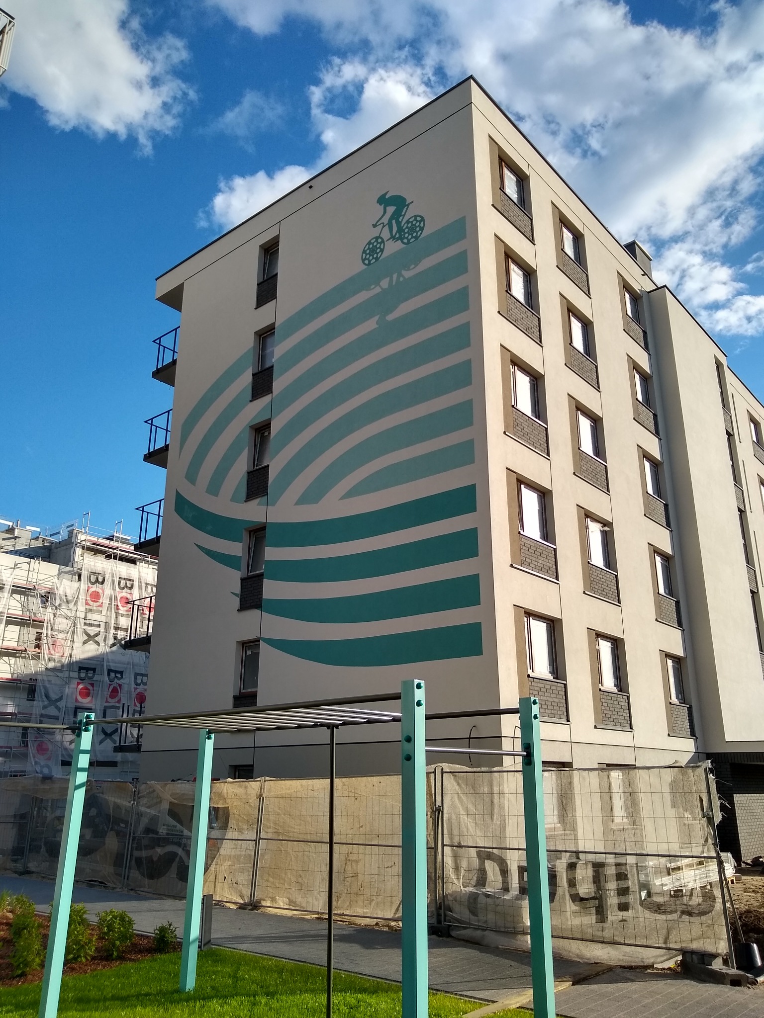 Wielopiętrowy budynek z nowoczesną elewacją, ozdobiony artystycznym malunkiem przedstawiającym rowerzystę i abstrakcyjne turkusowe pasy, widoczny z poziomu gruntu, z elementami placu budowy w tle.