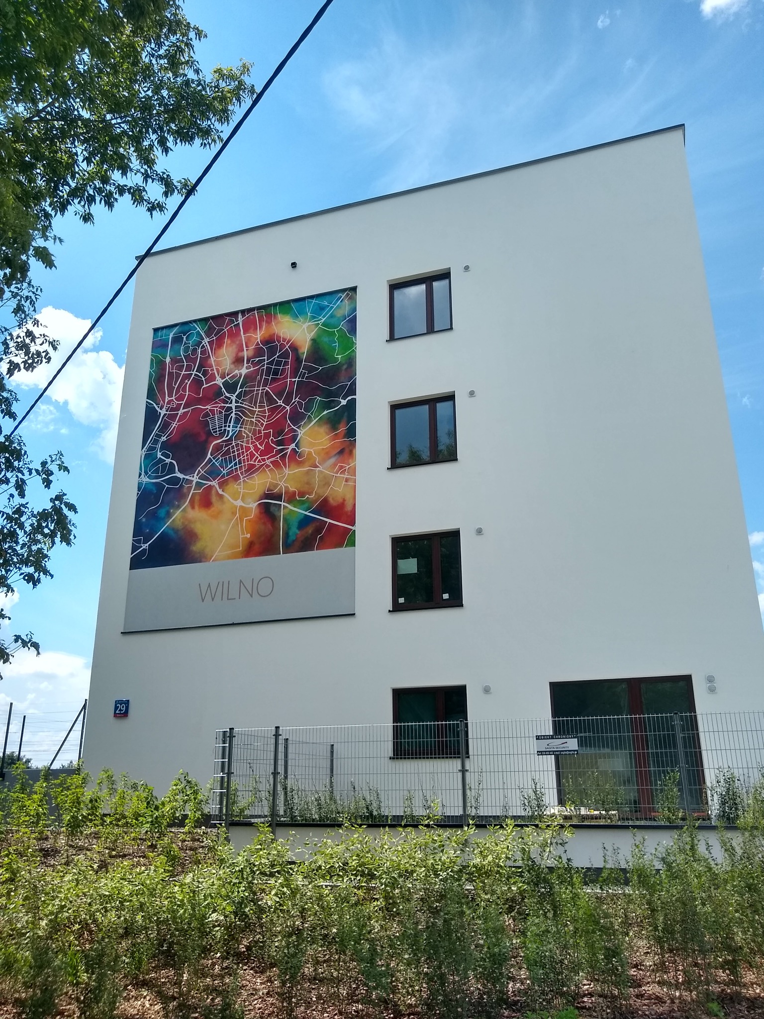 Biały budynek z dużym, kolorowym muralem przedstawiającym mapę miasta Wilno, widoczne okna z ciemnymi ramami i numer budynku 29.