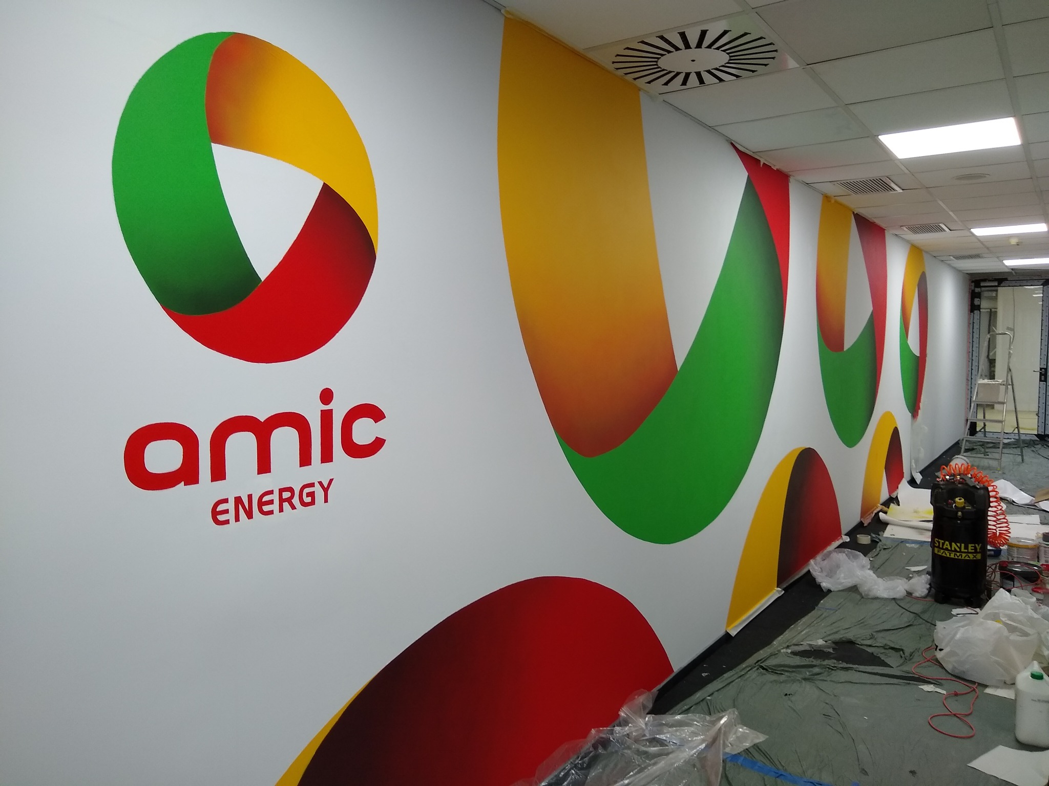 Naklejanie folii z logo Amic Energy na ścianę w biurze, widoczny kompresor Stanley Fatmax i narzędzia pracy.
