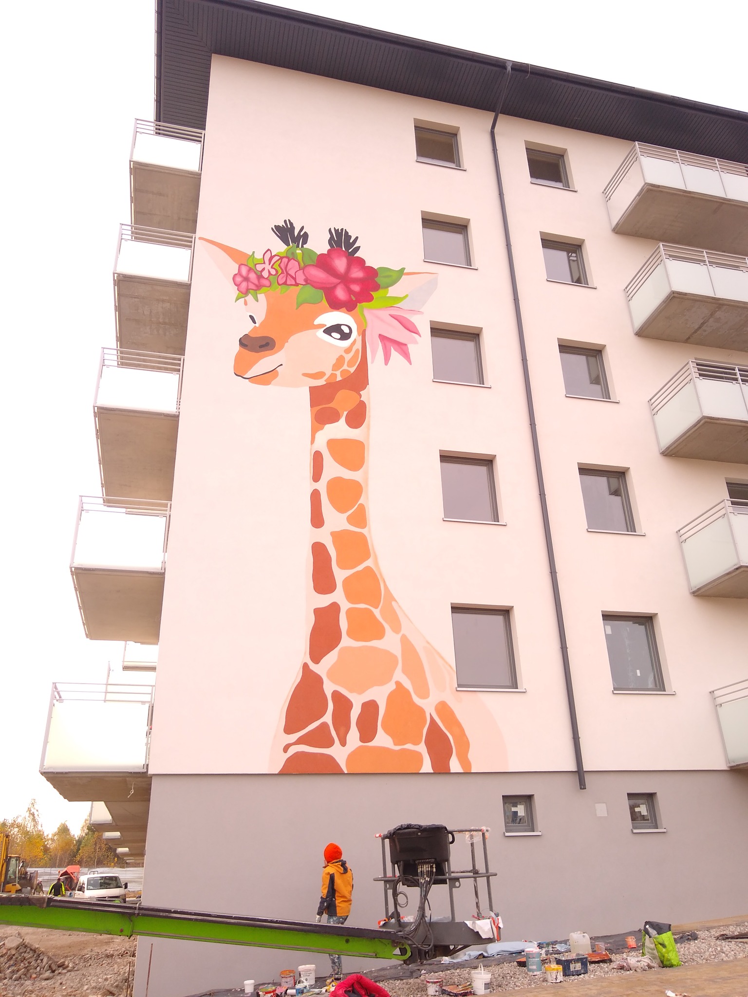 Świeżo ukończony mural na ścianie budynku mieszkalnego przedstawiający żyrafę z wiankiem z kwiatów na głowie, malowany z podnośnika przez osobę w pomarańczowej kurtce, widoczne farby i narzędzia.
