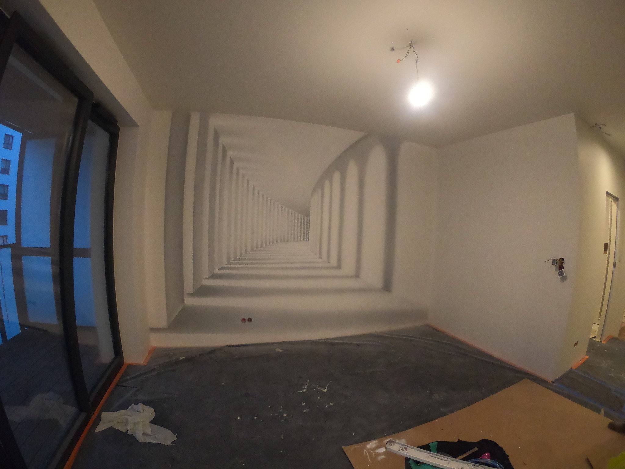 Wnętrze pokoju w trakcie prac: na ścianie widoczne malowidło iluzjonistyczne przedstawiające kolumnadę, podłoga zabezpieczona folią malarską, widoczne narzędzia malarskie i poziomica.