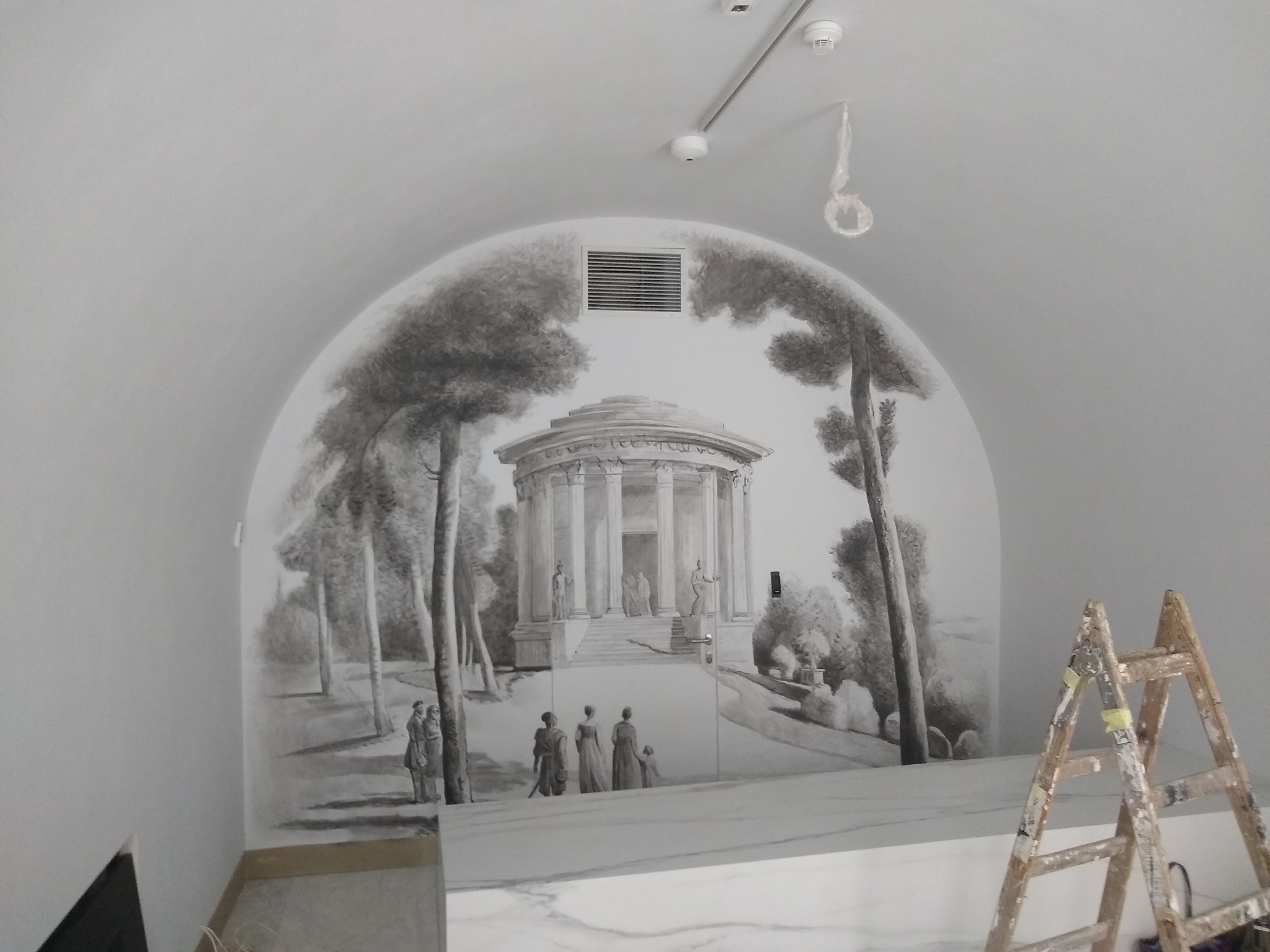 Ściana z ręcznie malowaną, szaro-białą iluzją optyczną przedstawiającą klasycystyczną świątynię i spacerujących ludzi, drabina malarska z boku.