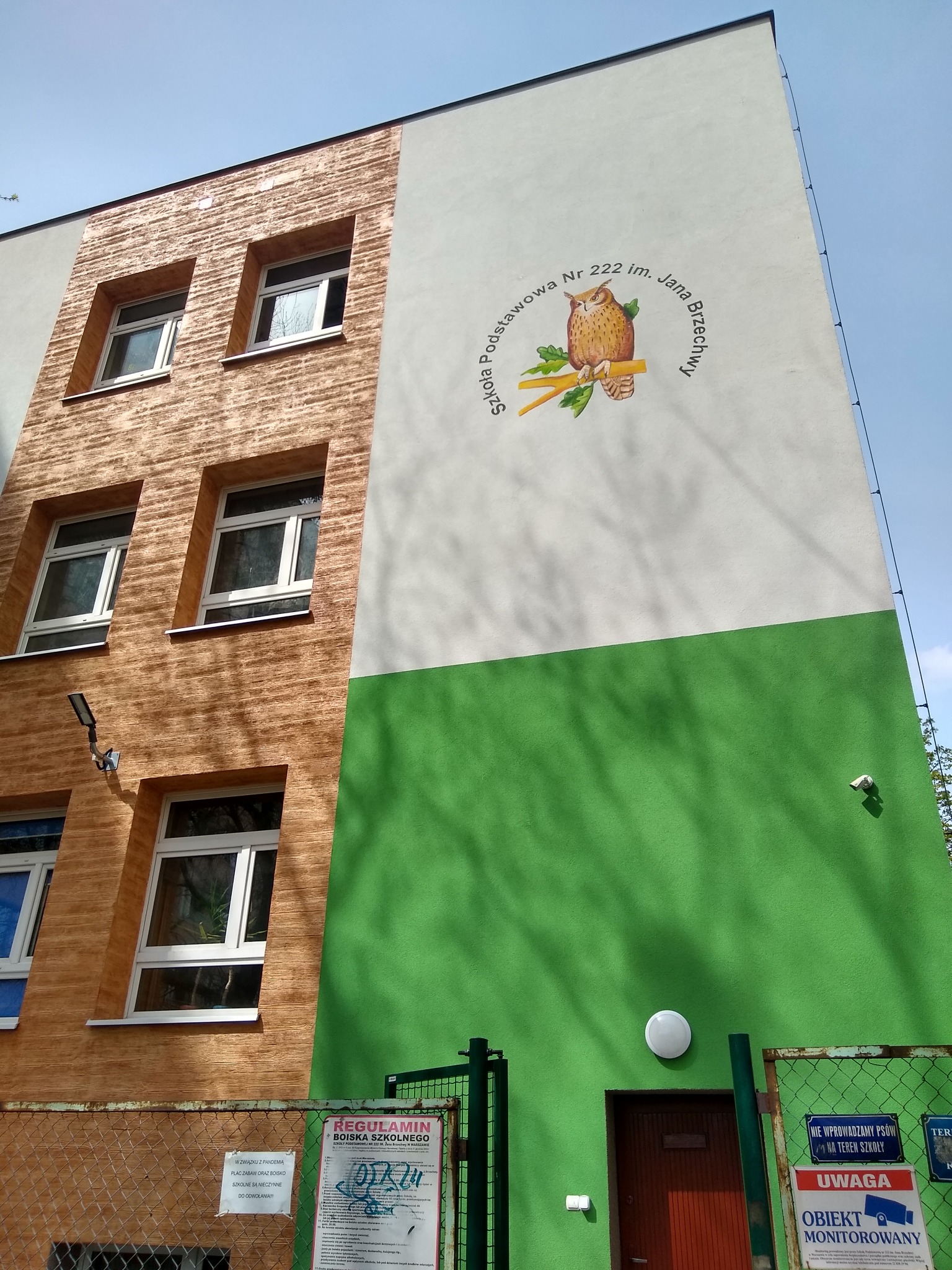 Odnowiona elewacja budynku szkoły podstawowej z brązową teksturą imitującą drewno, białym pasem z wizerunkiem sowy i zielonym dołem, widok z dołu na budynek z bramą i ogrodzeniem.
