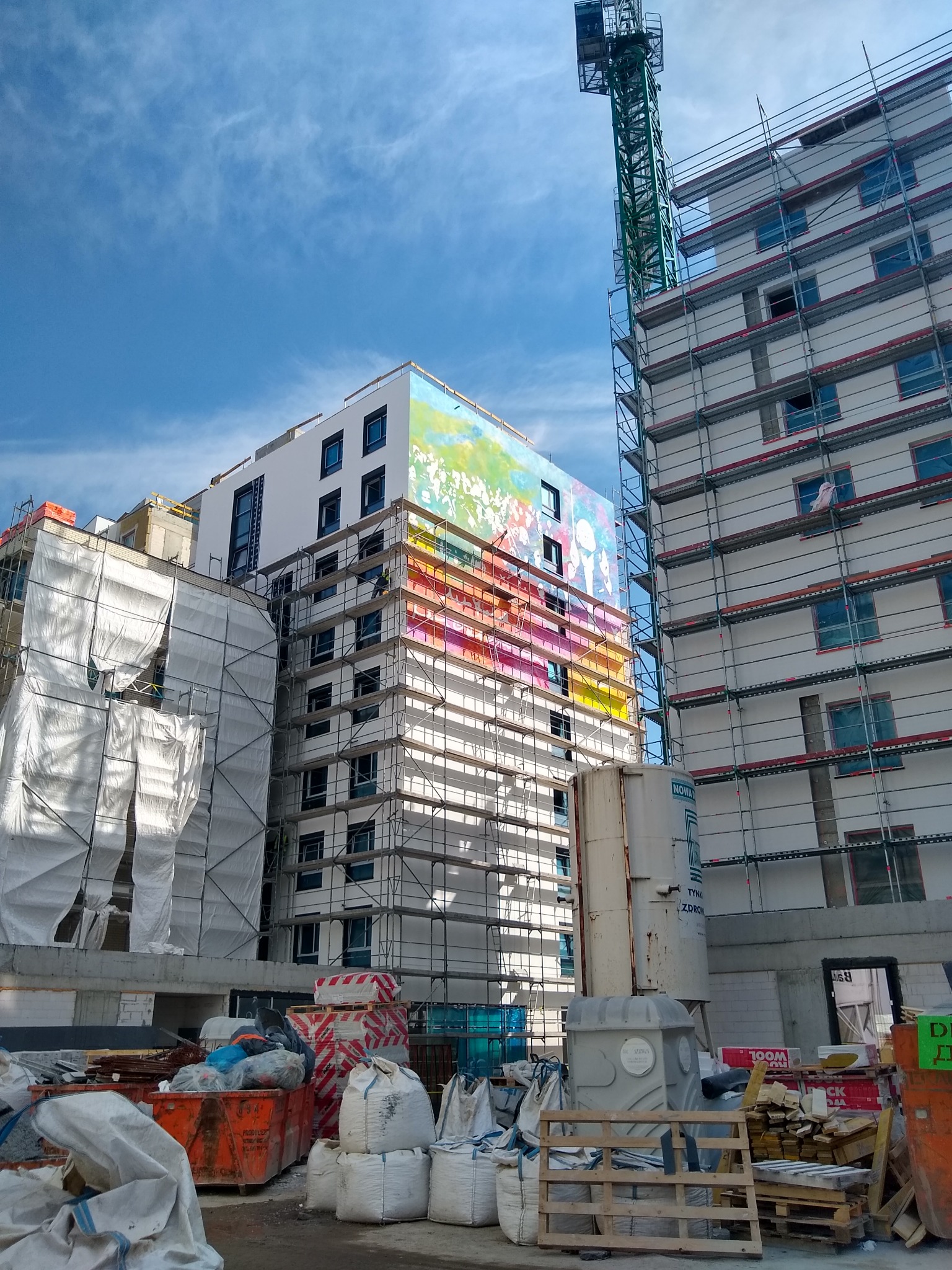 Wielopiętrowy budynek w trakcie budowy, z rusztowaniami i dźwigiem, elewacja częściowo pokryta kolorową abstrakcyjną grafiką, widoczne materiały budowlane i pojemniki na odpady.