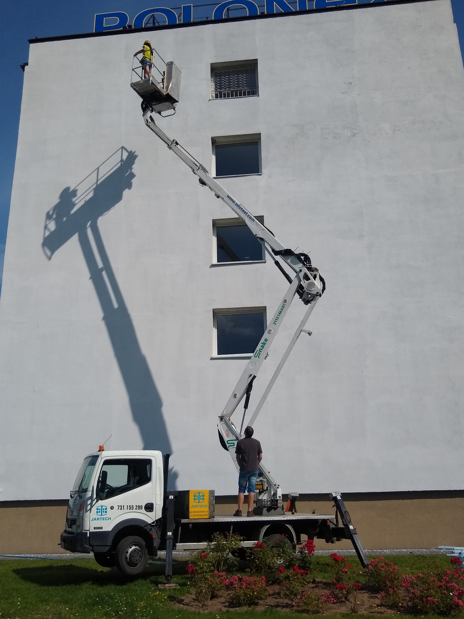 Pracownik w podnośniku koszowym na tle białej elewacji budynku z napisem, cień podnośnika na ścianie, na dole samochód firmy Jartech i rabata z różami.