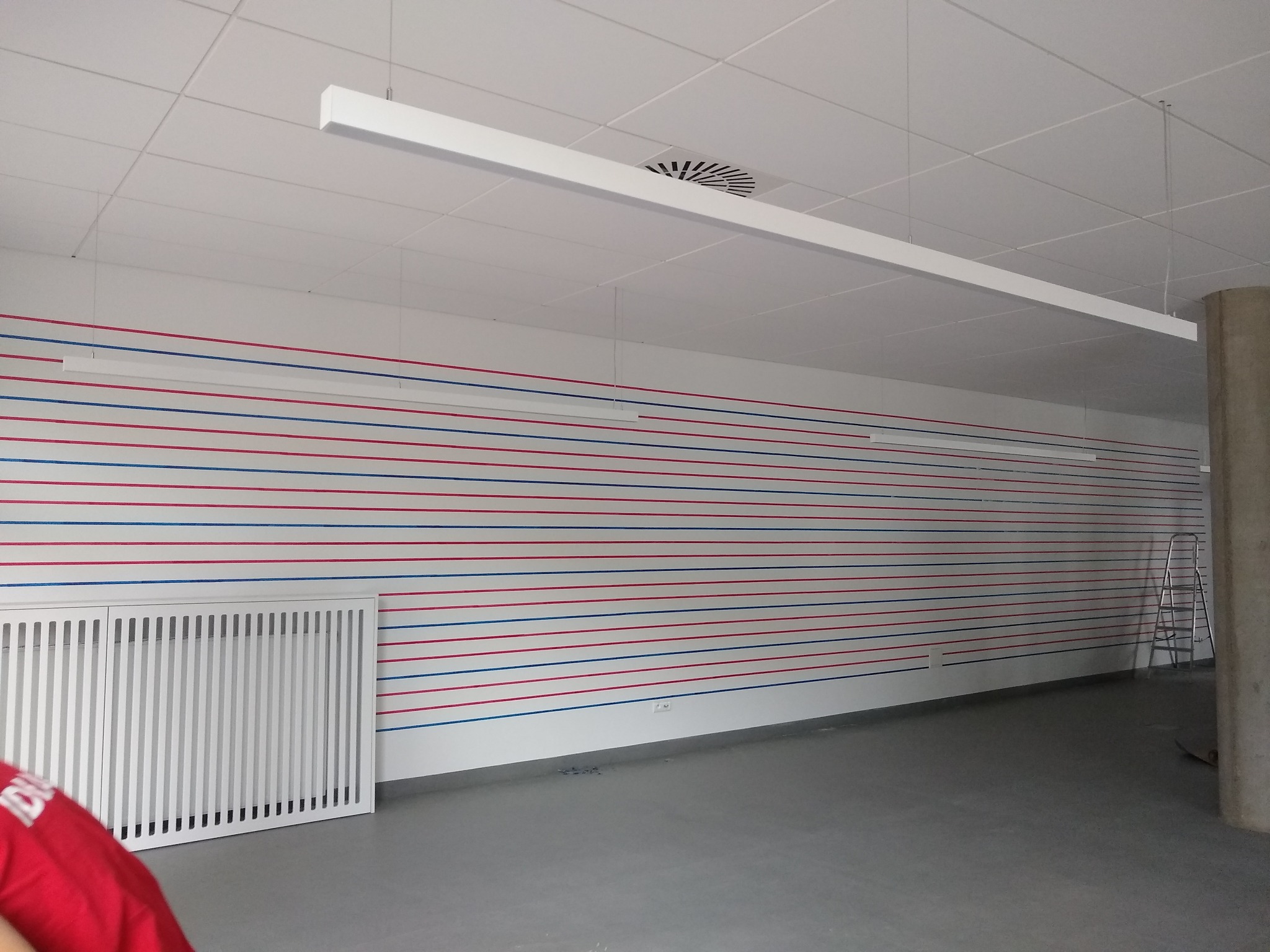 Wnętrze biura z pomalowaną ścianą w poziome, naprzemienne pasy koloru czerwonego i niebieskiego. Widoczny biały grzejnik, sufit kasetonowy z lampą liniową i kratką wentylacyjną oraz drabina oparta...