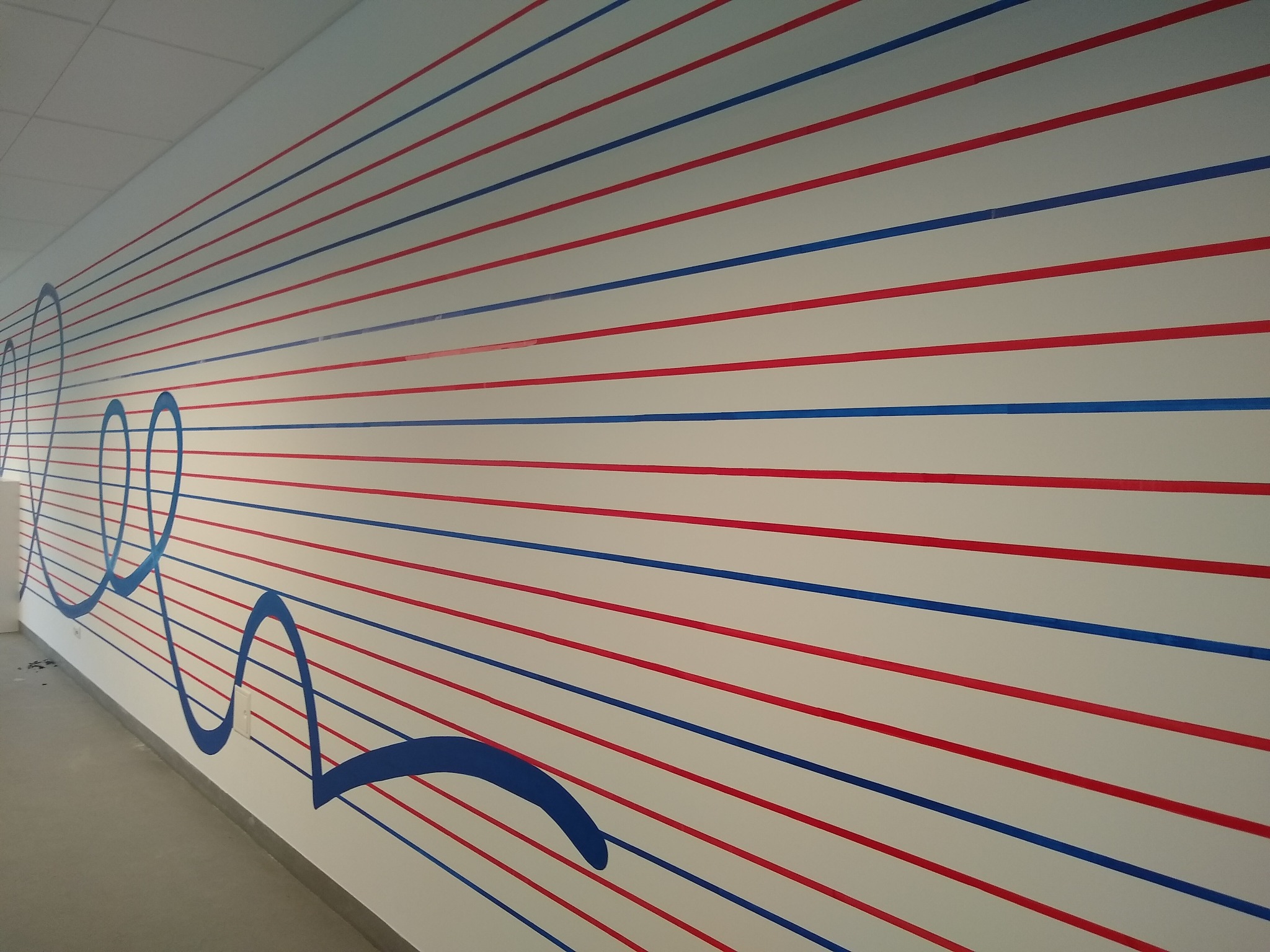 Ściana w korytarzu z poziomymi pasami w kolorach czerwonym i niebieskim oraz abstrakcyjnymi, zaokrąglonymi kształtami w kolorze niebieskim, przypominająca pięciolinię.