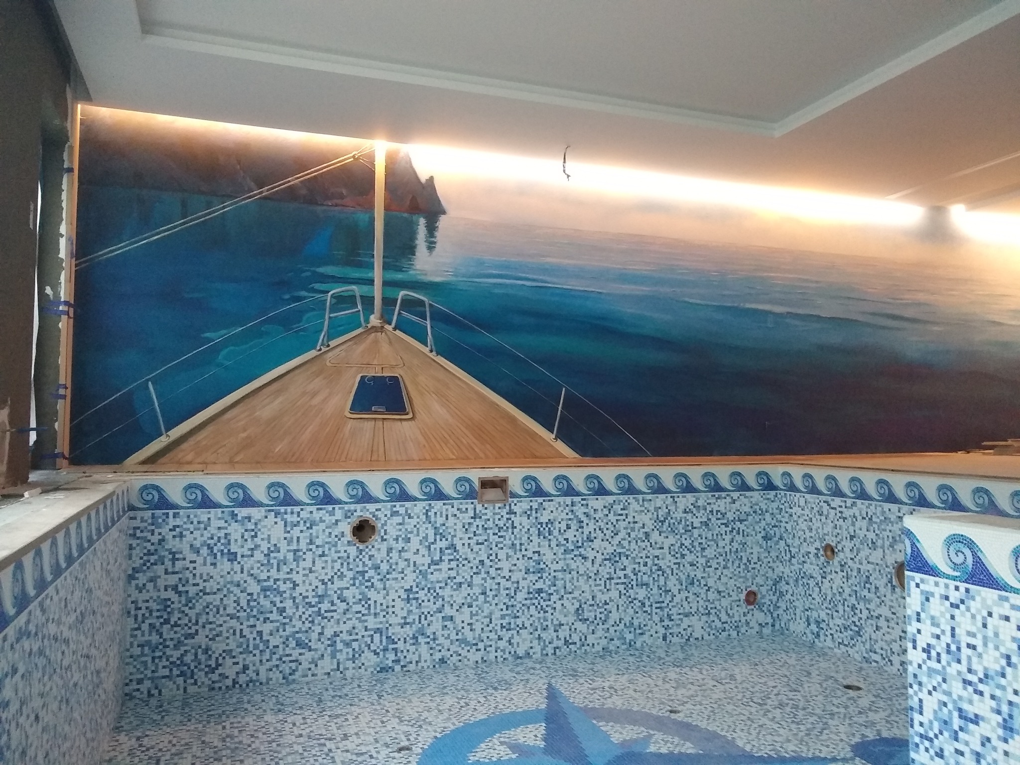 Niedokończony basen z mozaiką w odcieniach błękitu i bieli, z dekoracyjnym motywem fal na krawędziach i wzorem kompasu na dnie, uzupełniony malowidłem ściennym przedstawiającym łódź na tle...