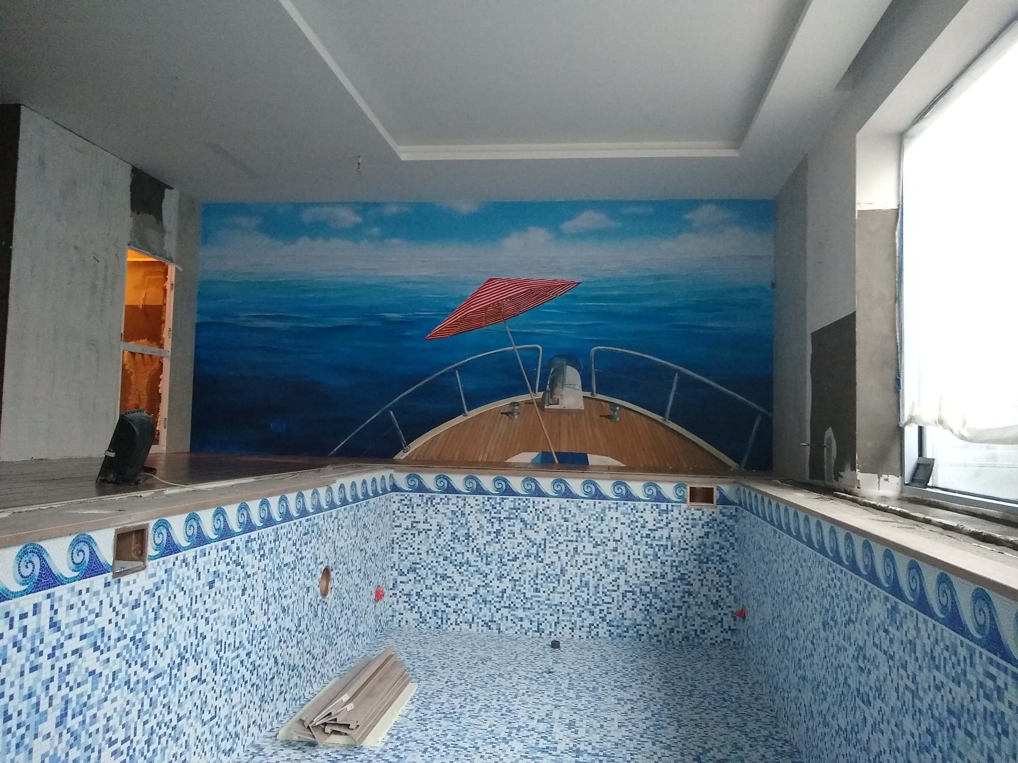 Wnętrze basenu w trakcie wykańczania, z mozaiką w odcieniach błękitu i dekoracyjnym fryzem z motywem fal, na ścianie mural z motywem morza i łodzi z czerwonym parasolem.