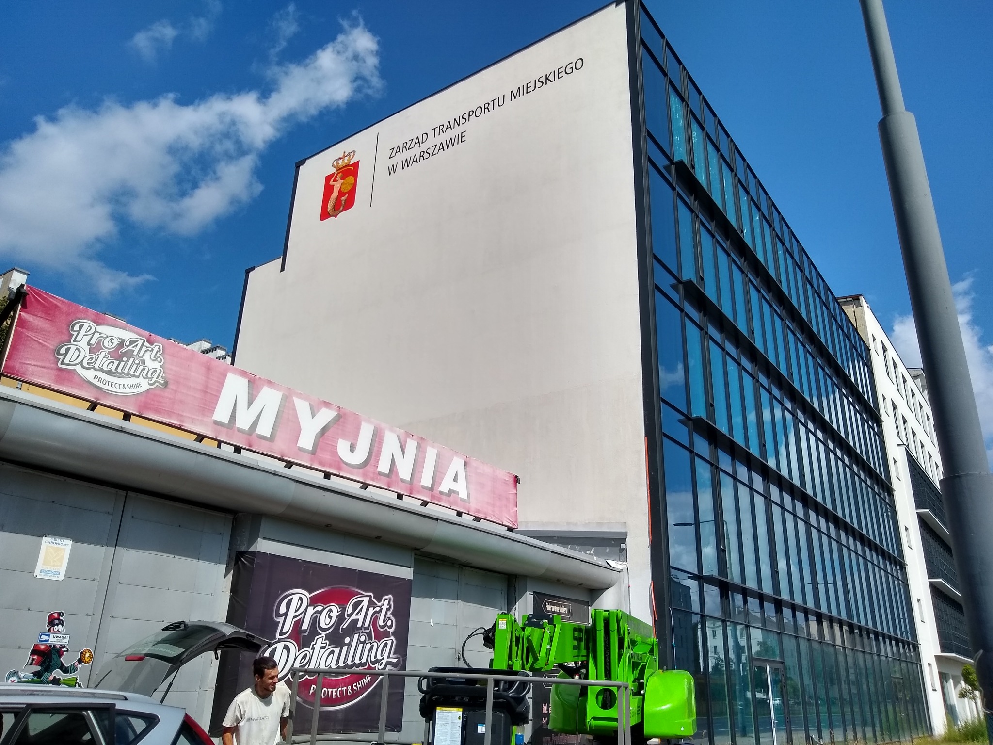 Budynek Zarządu Transportu Miejskiego w Warszawie z herbem miasta na elewacji, obok myjnia samochodowa z napisem 'MYJNIA' i banerami reklamowymi Pro Art Detailing.