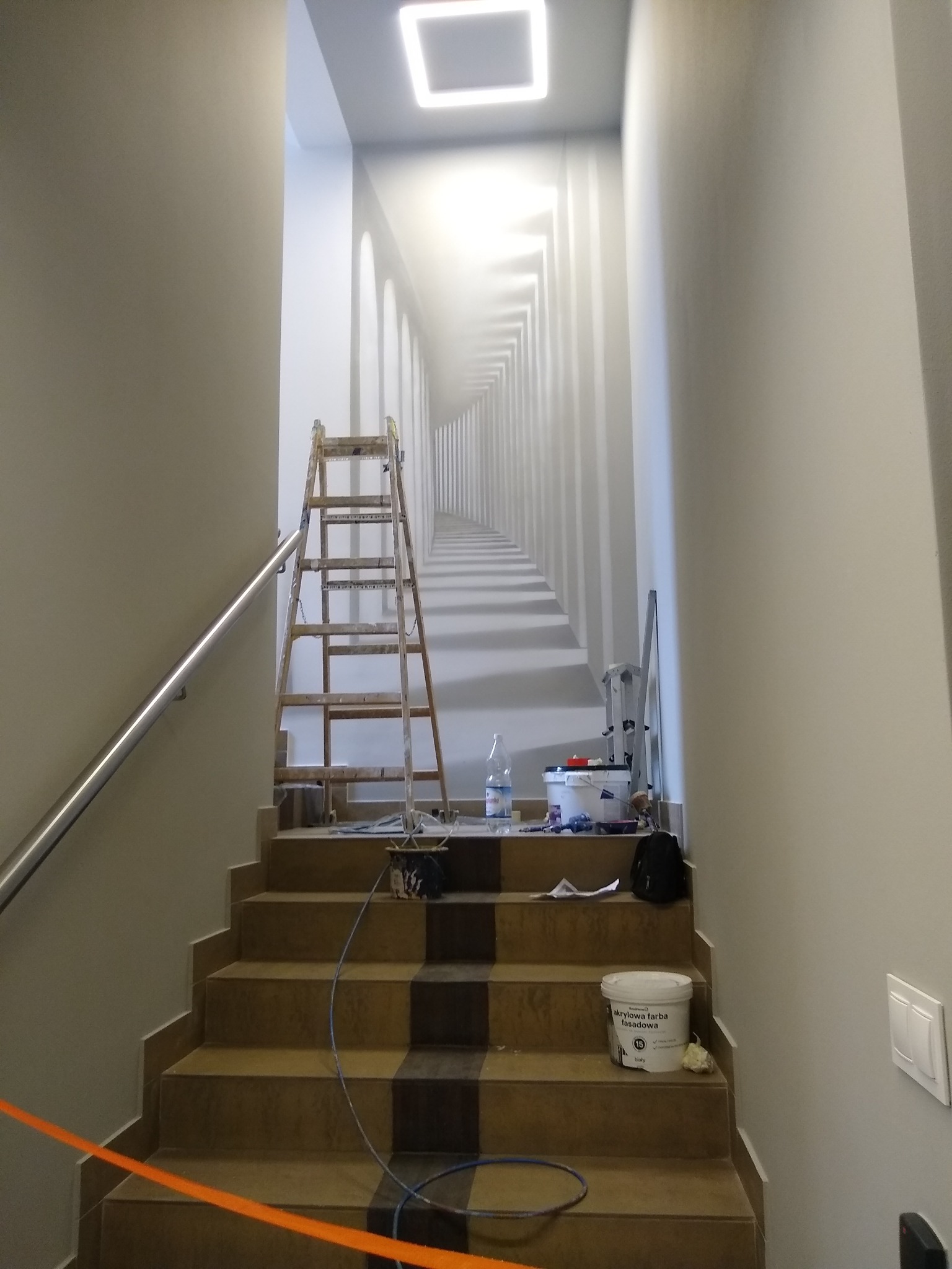 Klatka schodowa w trakcie odświeżania: drabina oparta o ścianę z fototapetą imitującą perspektywę kolumnady, wiadro z białą farbą akrylową na schodach, narzędzia malarskie i butelka wody w pobliżu.