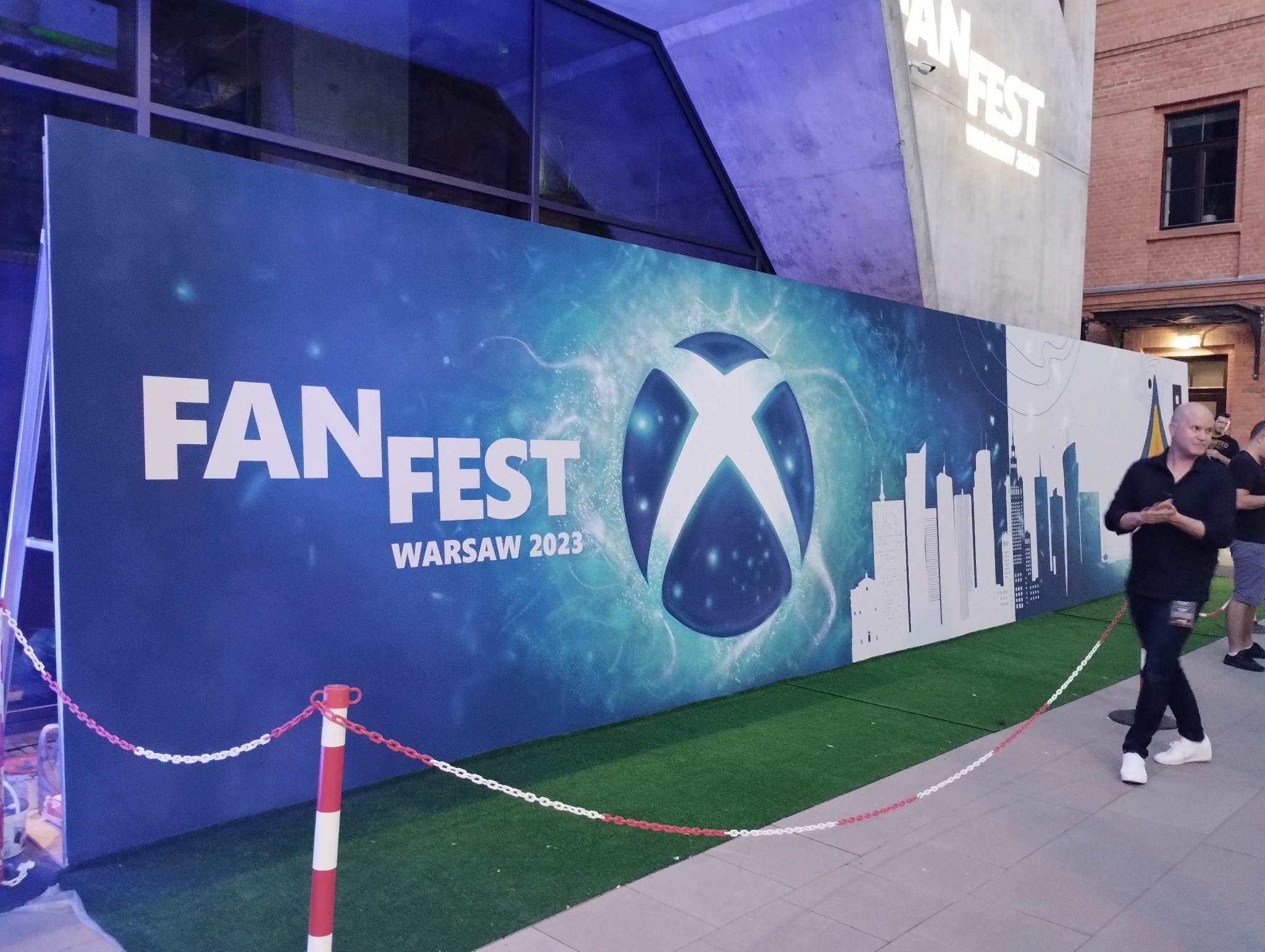 Baner reklamowy wydarzenia Fan Fest Warsaw 2023 z logo Xbox i sylwetką Warszawy, ustawiony na zewnątrz budynku, oddzielony łańcuchem od przechodniów.