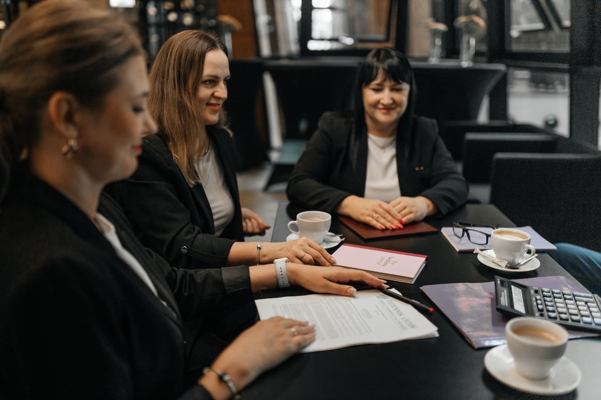 Trzy kobiety w biurowych strojach omawiają dokumenty przy stole z kawą i kalkulatorem. Spotkanie biznesowe w nowoczesnym wnętrzu.