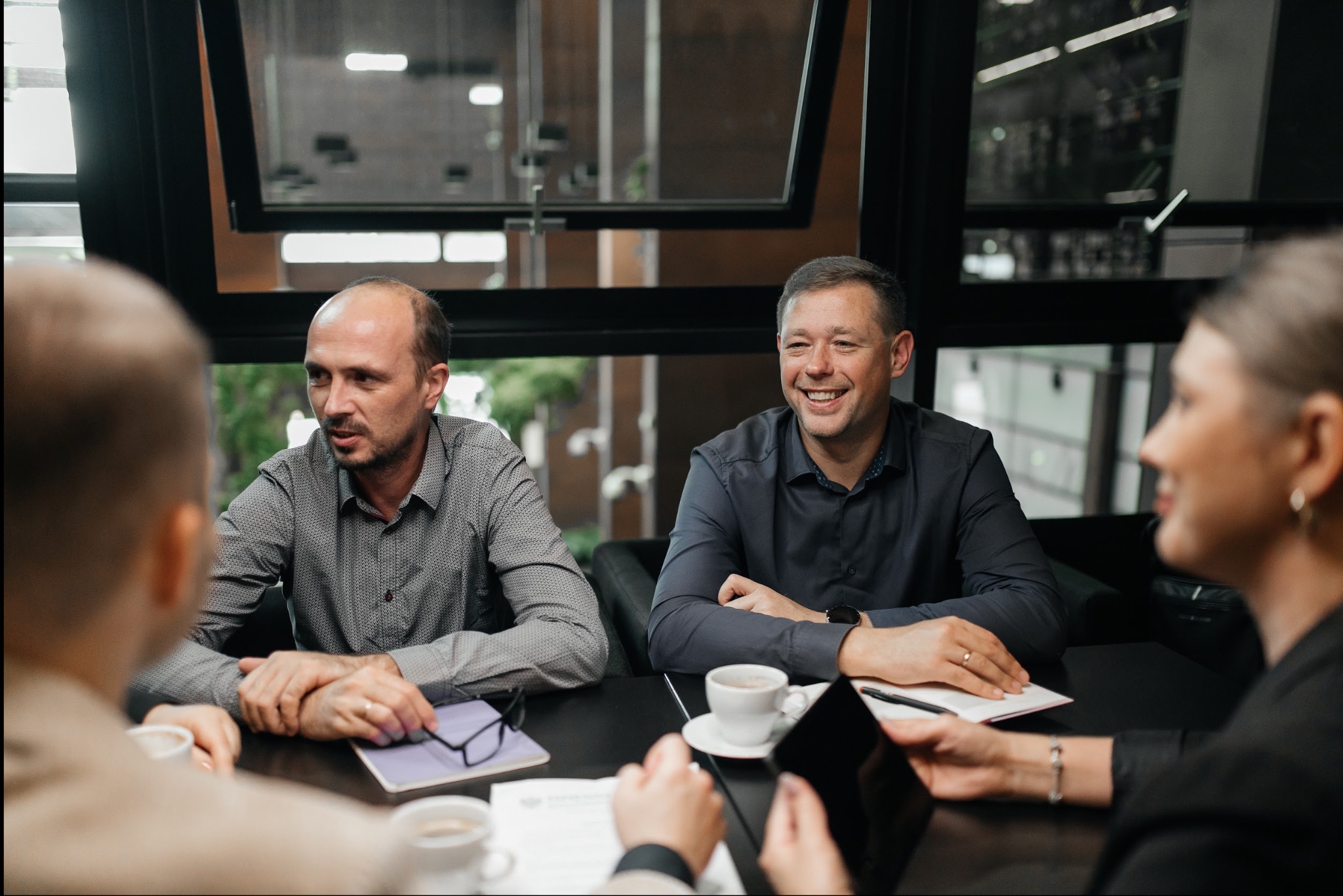 Spotkanie biznesowe w nowoczesnym biurze: mężczyźni i kobieta przy stole, notatki, kawa, naturalne światło przez okno. Jeden z mężczyzn uśmiecha się.