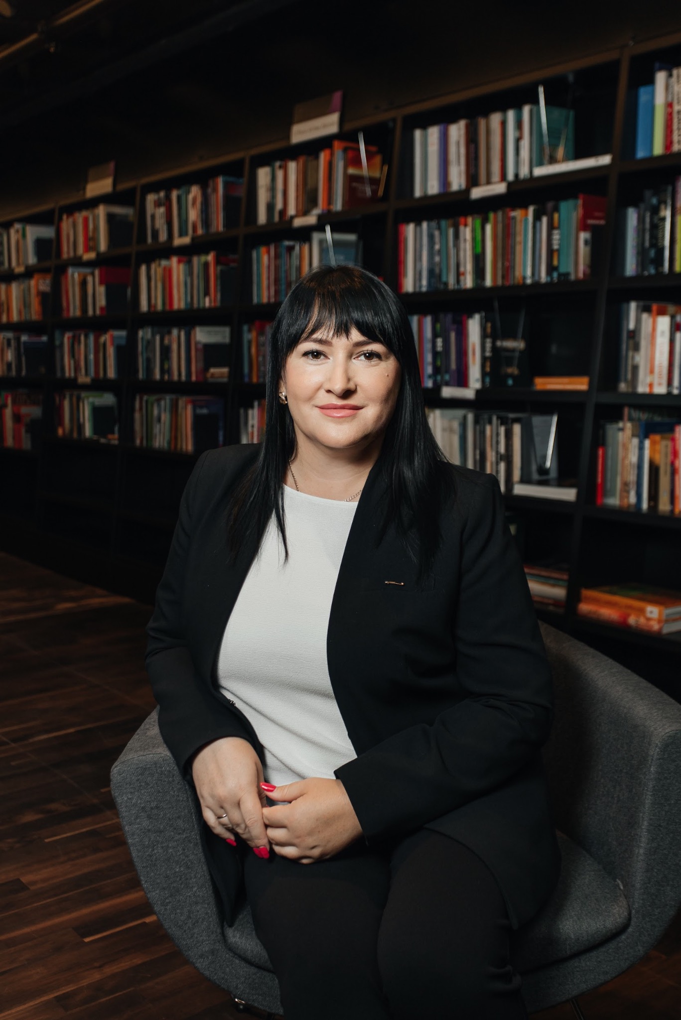 Kobieta w czarnym żakiecie siedzi na fotelu, za nią regał z książkami. Uśmiechnięta, patrzy w obiektyw. Profesjonalny wizerunek.