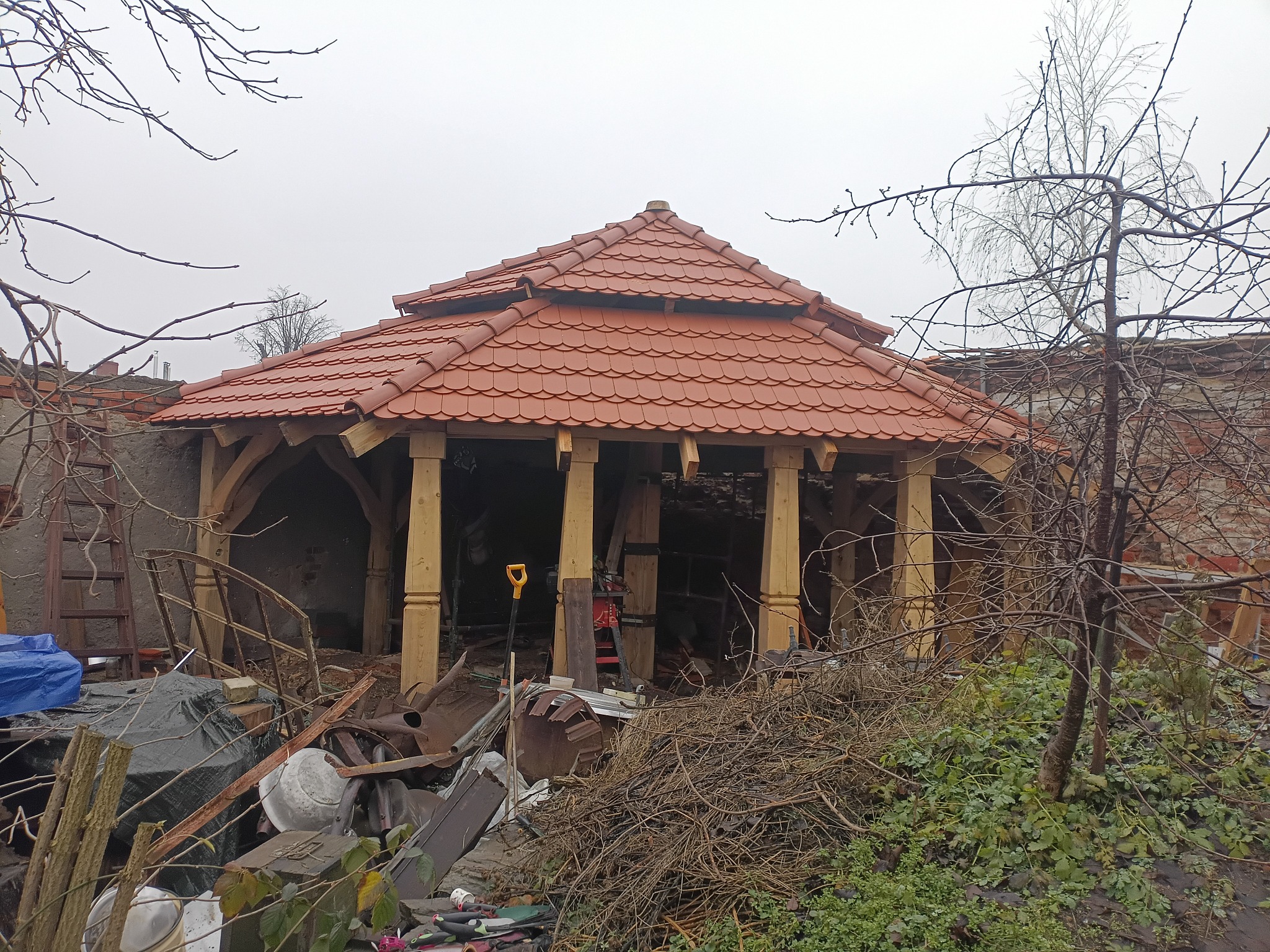 Drewniana altana z czerwoną dachówką w trakcie budowy, widoczne rusztowania i narzędzia. Surowy krajobraz, pochmurne niebo. Żółta łopata oparta o konstrukcję.