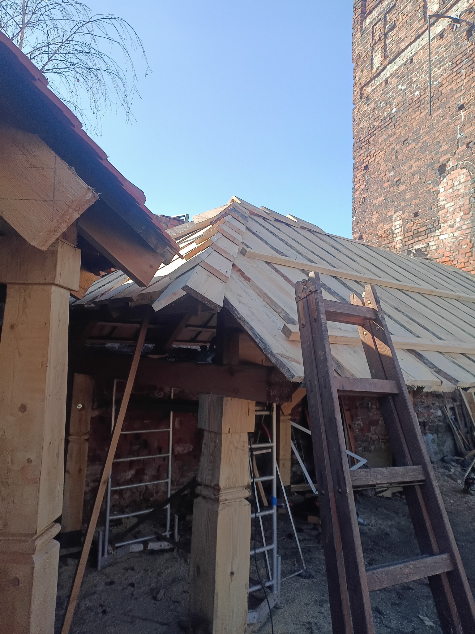 Konstrukcja dachu w trakcie budowy z widocznymi łatami i elementami drewnianymi, drabina oparta o ścianę z cegły w tle, jasne niebo.