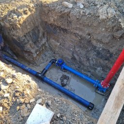 INSTAL-ART S.C. T. KLOC, S. DUDEK. - Wykop z ułożonymi niebieskimi rurami wodociągowymi z łącznikami, częściowo zanurzonymi w wodzie, widoczny fragment czerwonej rury pionowej, ziemia z kamieniami i łopata w tle.