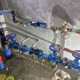 INSTAL-ART S.C. T. KLOC, S. DUDEK. - Kompleksowa instalacja wodociągowa z licznymi zaworami i rurami w kolorze niebieskim, zamontowana na surowej ścianie w trakcie budowy, widoczne narzędzia i poziomica.