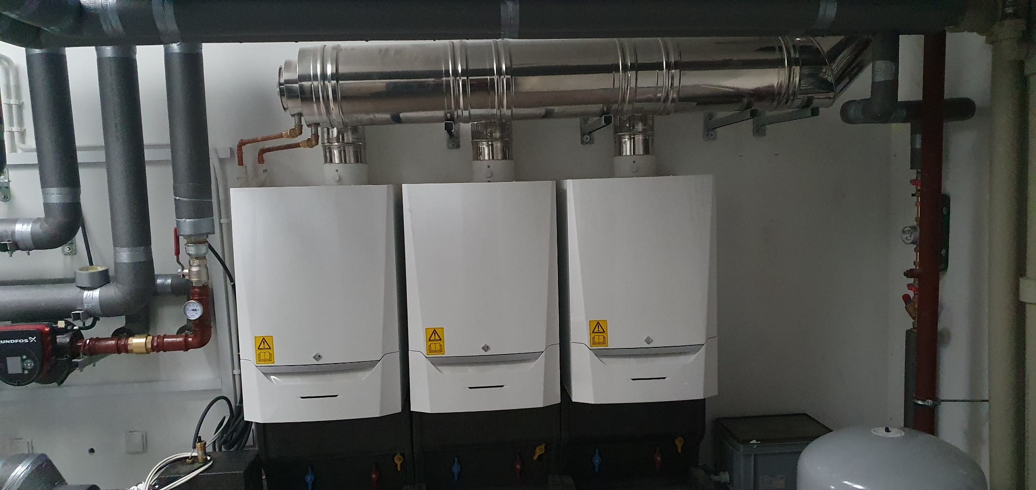 Trzy białe, nowoczesne kotły grzewcze zamontowane w kotłowni, z widocznymi rurami odprowadzającymi spaliny i instalacją hydrauliczną. Na kotłach naklejki ostrzegawcze.