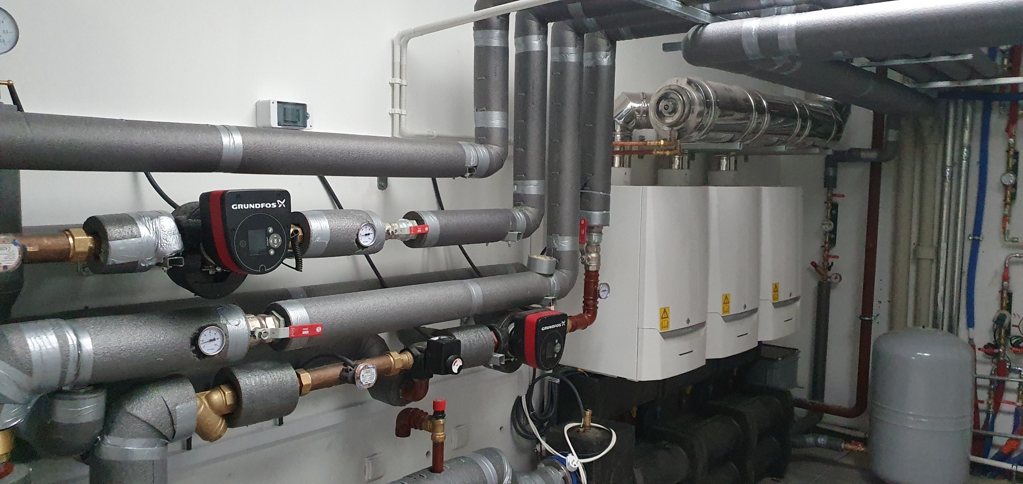 Instalacja grzewcza z izolowanymi rurami, pompami obiegowymi Grundfos i trzema białymi kotłami grzewczymi zamontowanymi na ścianie w kotłowni.