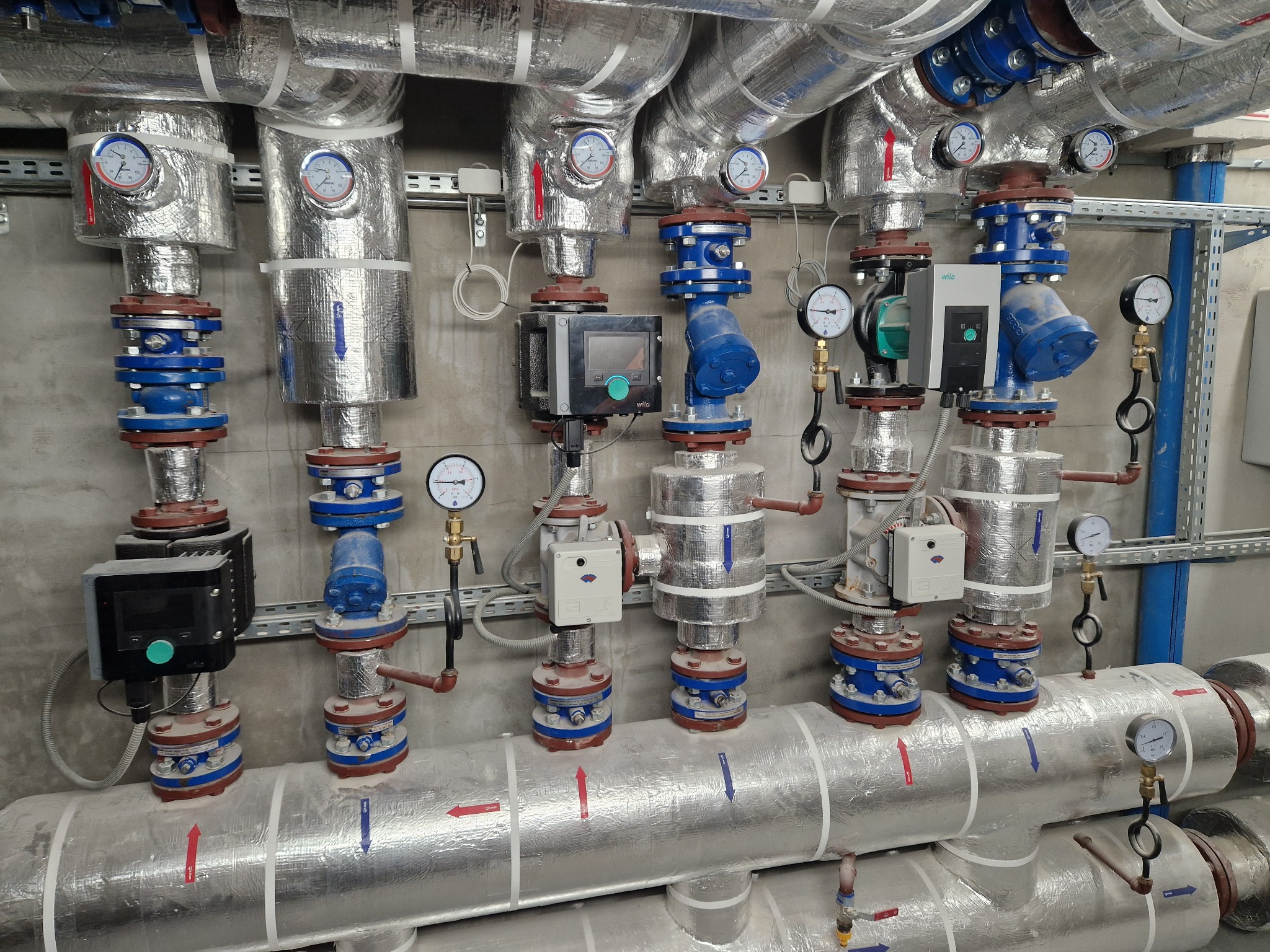 Izolowane rury instalacji grzewczej z manometrami, zaworami i pompami obiegowymi w kotłowni, widoczne strzałki wskazujące kierunek przepływu.