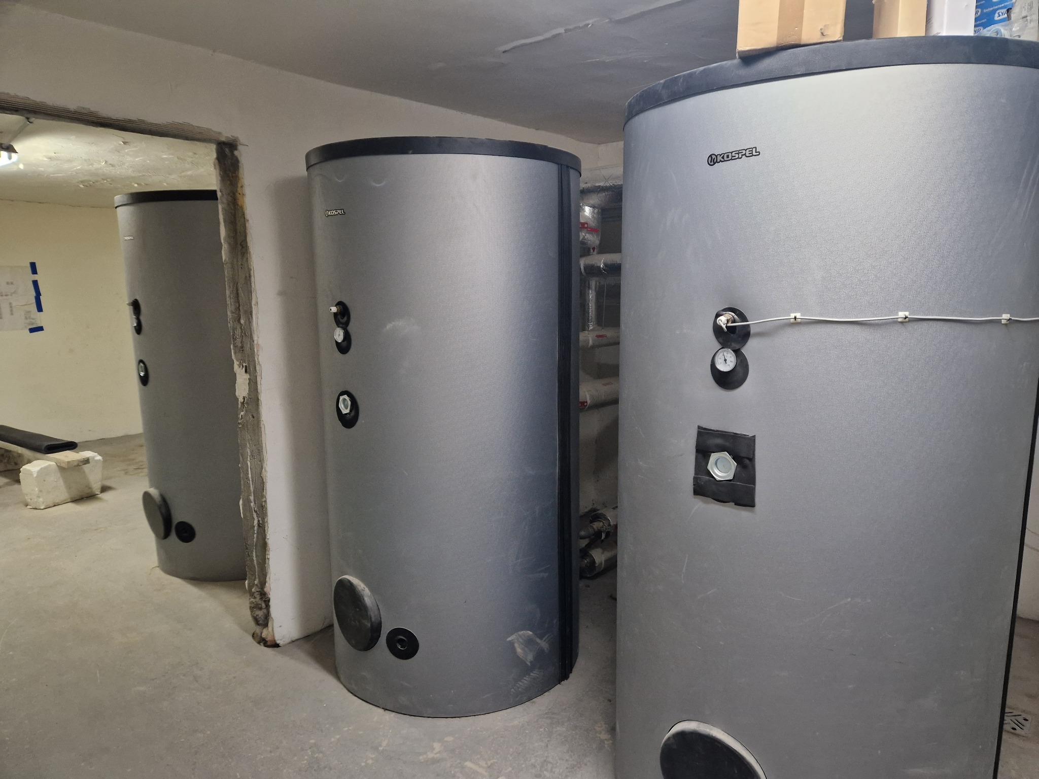 Szare, cylindryczne zbiorniki akumulacyjne Kospel w piwnicy w Jankowicach, widoczne rury instalacyjne w tle.