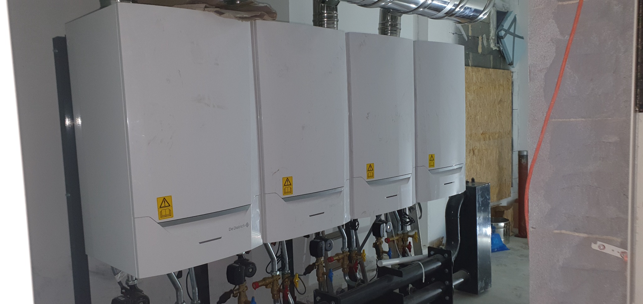 Cztery białe, kondensacyjne kotły grzewcze marki De Dietrich zamontowane na ścianie, z widocznymi rurami i zaworami w pomieszczeniu w trakcie wykańczania.