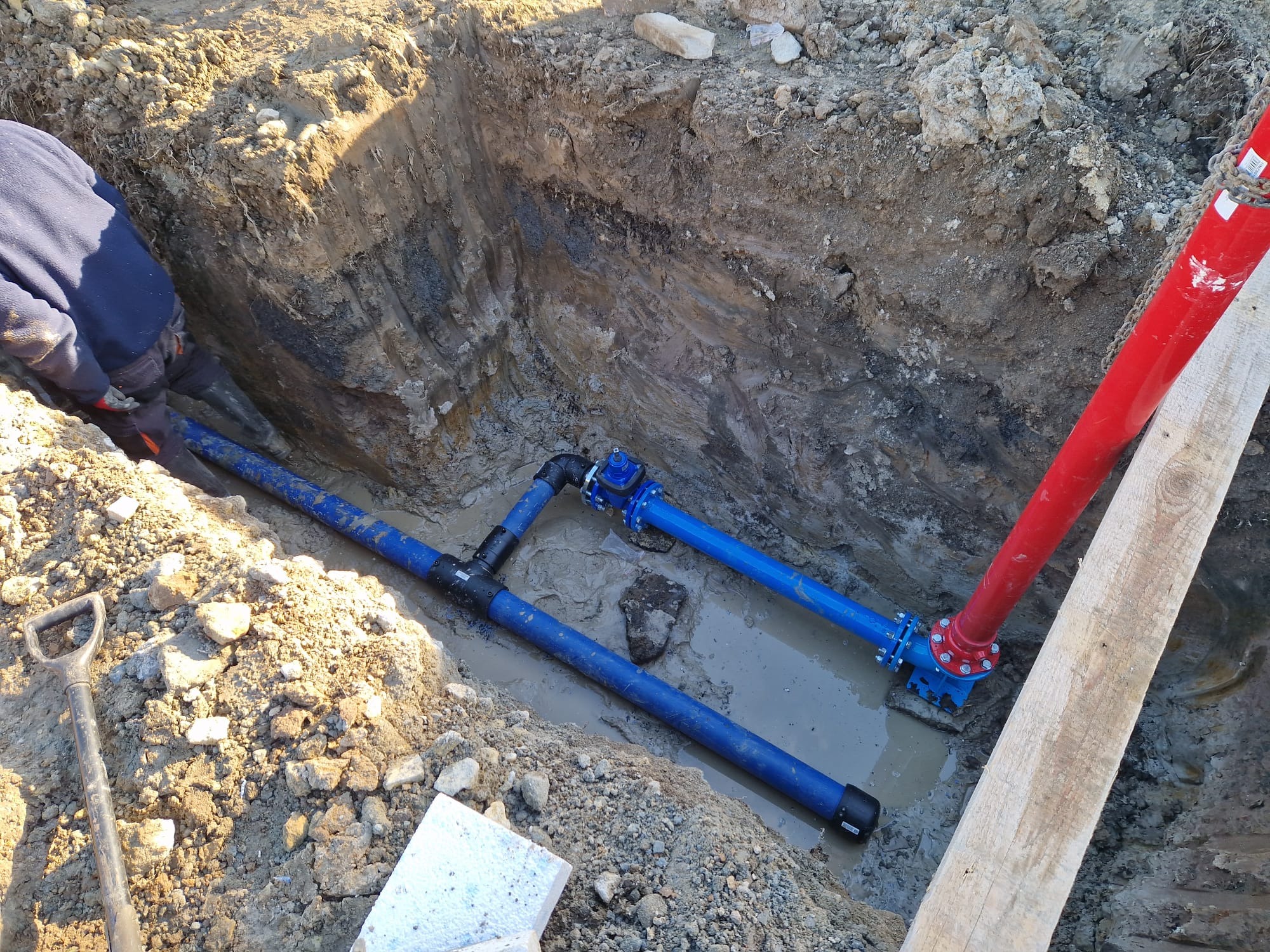 Wykop z ułożonymi niebieskimi rurami wodociągowymi z łącznikami, częściowo zanurzonymi w wodzie, widoczny fragment czerwonej rury pionowej, ziemia z kamieniami i łopata w tle.