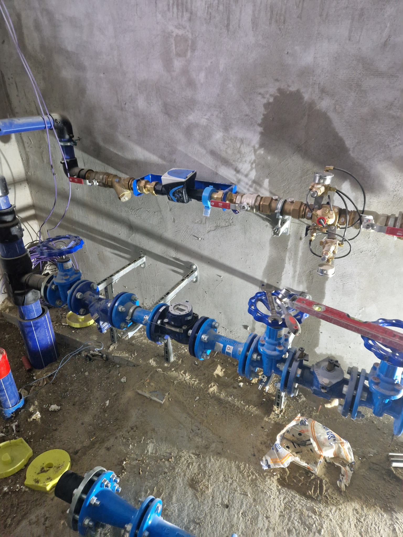 Kompleksowa instalacja wodociągowa z licznymi zaworami i rurami w kolorze niebieskim, zamontowana na surowej ścianie w trakcie budowy, widoczne narzędzia i poziomica.