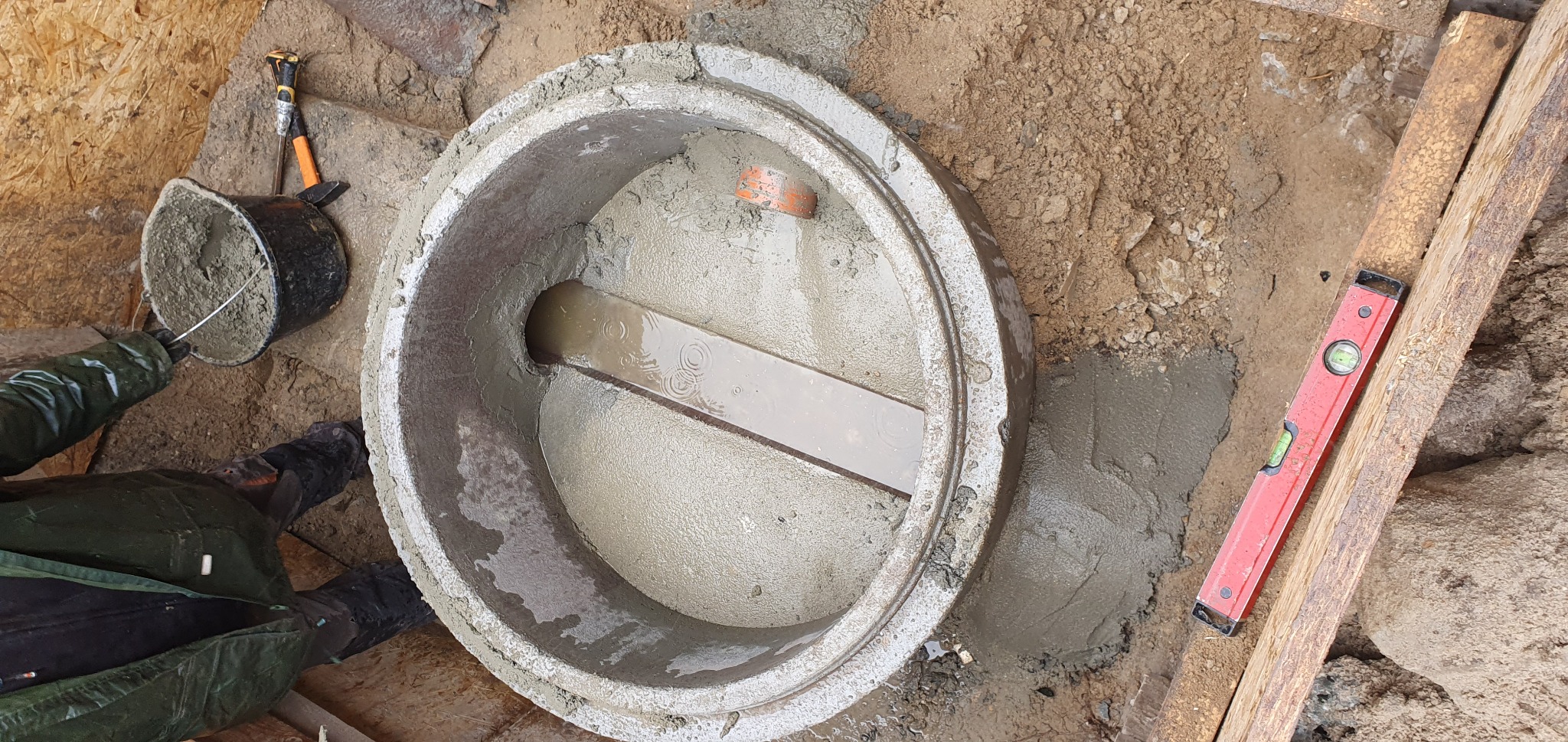 Montaż betonowego kręgu studziennego z widocznym dnem, częściowo wypełnionego wodą, z deską wewnątrz i poziomnicą obok, w trakcie prac ziemnych.