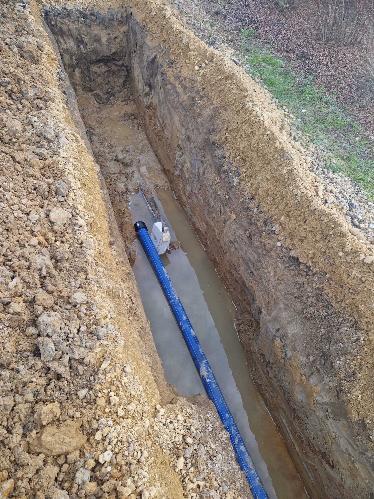 Wykop w ziemi wypełniony wodą, w którym ułożona jest niebieska rura, widoczne ślady po wykopie i pryzma ziemi obok.